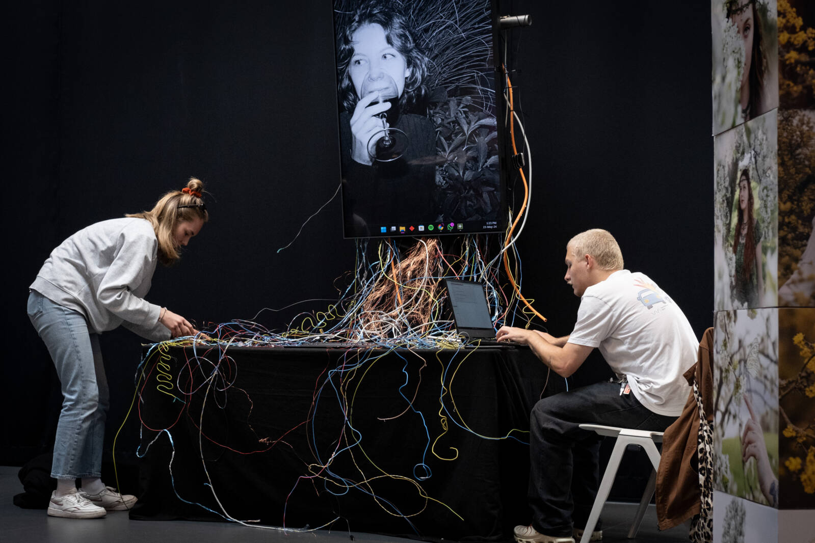 Zwei Personen arbeiten an einer Kunst- und Technologieinstallation mit bunten Kabeln, die zu einem großen Bildschirm mit einem Schwarz-Weiß-Porträt führen. Eine Person richtet die Kabel, während die andere am Laptop arbeitet. Zwei Personen arbeiten an einer Kunst- und Technologieinstallation mit bunten Kabeln, die zu einem großen Bildschirm mit einem Schwarz-Weiß-Porträt führen. Eine Person richtet die Kabel, während die andere am Laptop arbeitet.