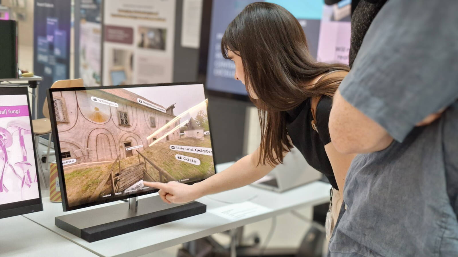 Eine Frau bedient einen Touchscreen mit einem 3DModell eines historischen Gebäudes, während eine weitere Person daneben zusieht. Eine Frau bedient einen Touchscreen mit einem 3DModell eines historischen Gebäudes, während eine weitere Person daneben zusieht.